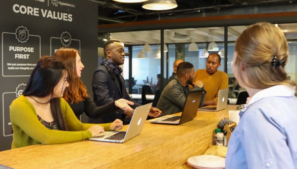 7 people (3 white female and four males, including three black ones, are sitting on a roundtable discussing. They are in HyperionDev office.