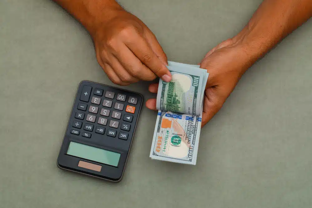 A photo of 100-dollar bills in the hands of a dark skinned person with a calculator on the side