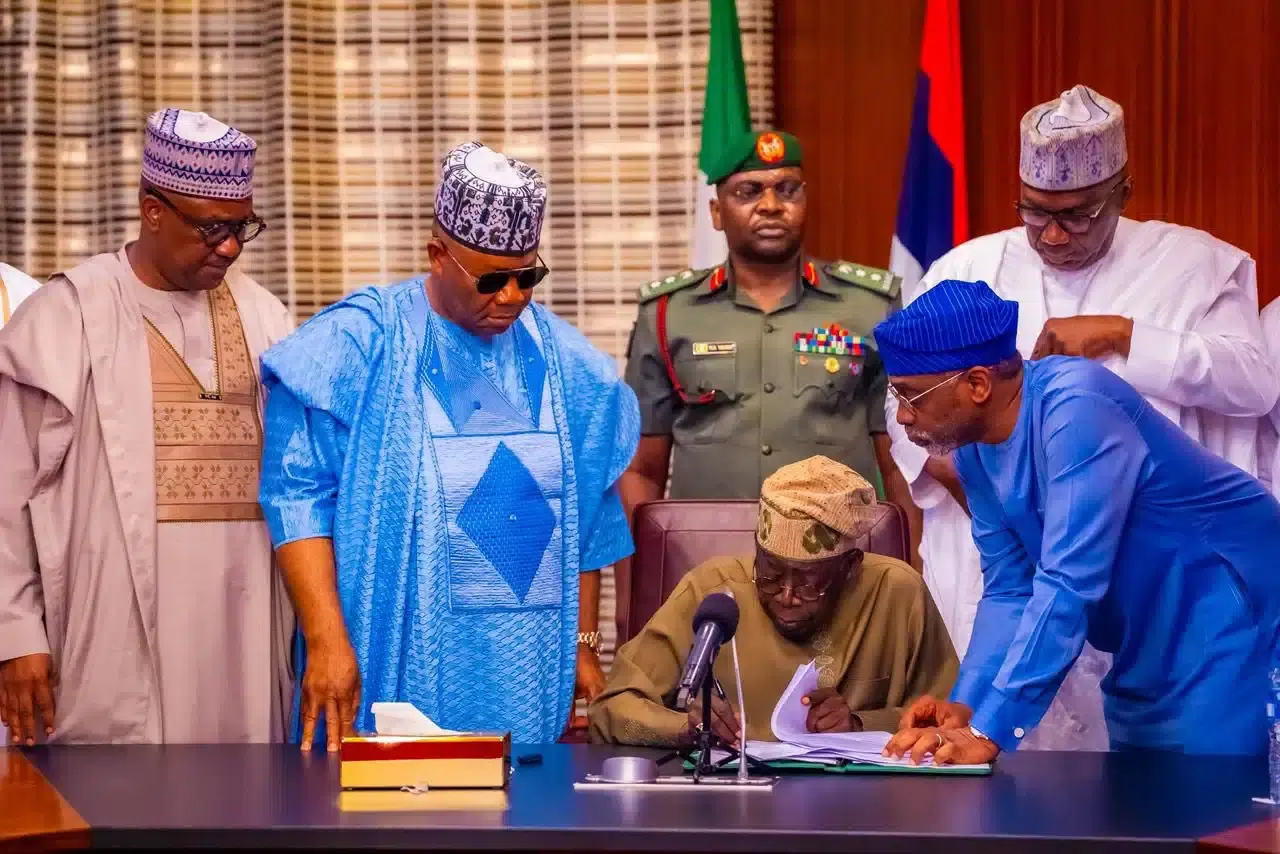 President Tinubu sits reviewing a document, while four men in agbada stand behind him, joined by another in military uniform.